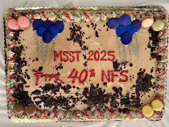 This is a festive chocolate celebration cake commemorating the 40th anniversary of NFS (Network File System). The rectangular cake has chocolate frosting as its base and is decorated with:
	•	Red piped text reading “MSST 2025” and “Happy 40th NFS”
	•	Colorful macarons arranged around the edges in various colors including blue, pink, green, and yellow
	•	Blue macarons arranged to look like balloons with green and white striped candy cane “strings”
	•	Chocolate cookie or cake crumbs scattered acr…