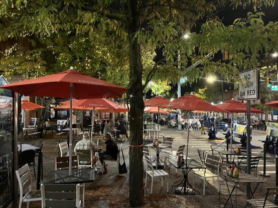 This is a vibrant evening scene of an outdoor dining area, part of a restaurant’s pedestrianized street dining setup. The space features multiple red umbrellas that create a cohesive canopy over the seating area, with modern metal chairs and small round tables arranged throughout.

The setting is a city street, with mature trees providing natural overhead coverage and string lights adding a warm, festive atmosphere. You can see several groups of diners enjoying their meals at various tables, cr…
