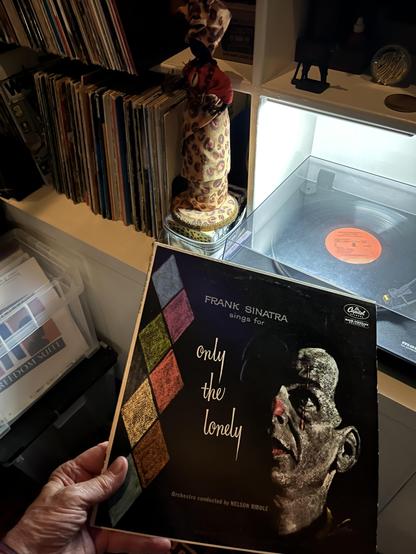 This photo shows a vinyl record collection and turntable setup. In the foreground, someone [hello] is holding a Frank Sinatra album titled "Only the Lonely" with "Sings for" visible on the cover, which features a black and white portrait of Sinatra. The album indicates it's conducted by Nelson Riddle and appears to be a Capitol Records release.

Behind the album, there's a turntable with its clear plastic cover open [what? it's closed], and you can see an orange/coral colored vinyl record on th…
