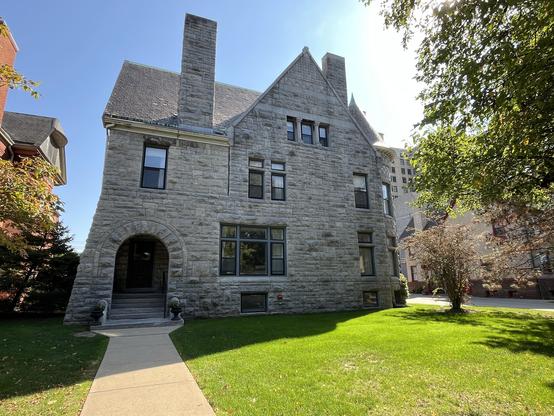 This photo shows a beautiful historic stone house with distinctive Romanesque Revival architecture. The building is constructed from gray limestone blocks, giving it a substantial, fortress-like appearance. Key architectural features include:
	•	A prominent arched entrance with thick stone framing
	•	Multiple tall chimneys rising from the slate or dark shingle roof
	•	A steep triangular gable with three grouped windows
	•	Various window styles throughout, including some with decorative stone tr…