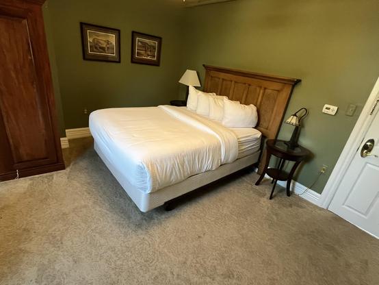 This photo shows a guest room with a warm, inviting atmosphere. The room features:

Bed and Furnishings:
	•	A queen-size bed with crisp white linens and multiple pillows
	•	A substantial wooden headboard with panel detailing in a dark wood finish
	•	A matching wooden nightstand with a table lamp featuring a white shade
Room Details:
	•	Sage green or olive-colored walls that create a calming ambiance
	•	Beige/tan carpeting throughout
	•	Two framed artwork pieces hanging on the wall above the bed…