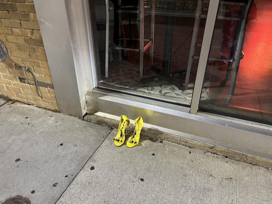 The photo shows a pair of bright yellow high-heeled sandals placed neatly on a sidewalk, right next to a building with a glass window and metal frame. The shoes are positioned side by side, facing outward toward the street. The sidewalk is concrete with some dark spots and marks, and the building has a brick wall on the left side. Inside the glass window, you can see part of a table base, a chair, and patterned flooring, with red lighting reflecting from inside. The scene appears to be taken at…
