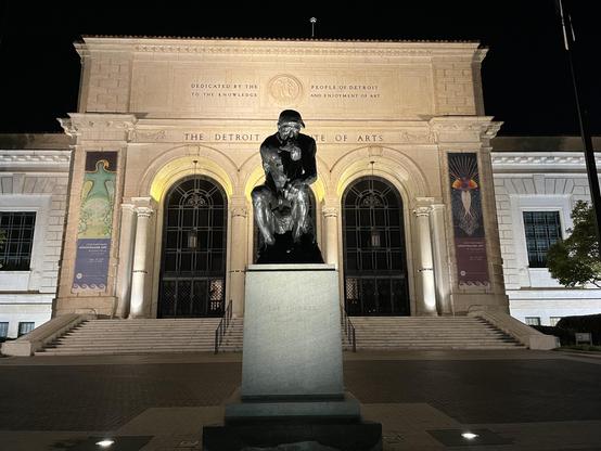 This is a nighttime photograph of the Detroit Institute of Arts, a grand neoclassical building with beautiful limestone or marble architecture. The facade features elegant columns, arched windows with decorative ironwork, and classical detailing. The building’s inscription reads “DEDICATED BY THE PEOPLE OF DETROIT TO THE KNOWLEDGE AND ENJOYMENT OF ART” at the top, with “THE DETROIT INSTITUTE OF ARTS” prominently displayed below.

The most striking feature is the large bronze sculpture positione…