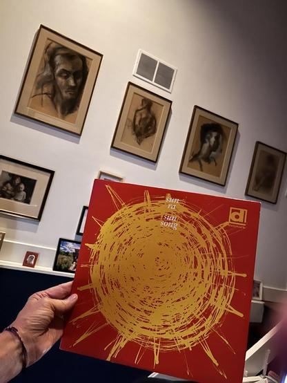 This photo shows someone [it absolutely me!] holding a vibrant red vinyl record album at home with framed artwork [by detroit artist Zubel Khachadoorian!] on the walls. The album cover features a striking gold circular design - a spiral or scribbled sun-like pattern with radiating lines extending outward like sun rays. The text on the album repeats "sun song" multiple times, and there's a small logo in the bottom right corner that appears to say "palmyra."

In the background, you can see severa…