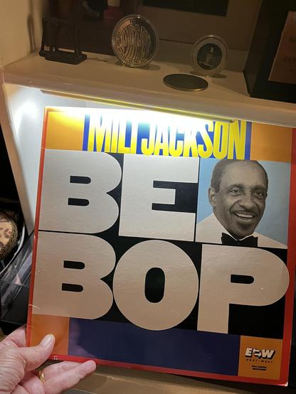 This photo shows someone [me again] holding a vibrant jazz album titled "BEBOP" by Milt Jackson. The cover features bold, eye-catching design with large white letters spelling "BEBOP" against a colorful background with orange, yellow, and blue sections. In the upper right corner is a photograph of Milt Jackson smiling, wearing formal attire with a bow tie. His name "MILT JACKSON" appears in yellow text at the top of the album.

The album is on the East-West label, with their logo visible in the…