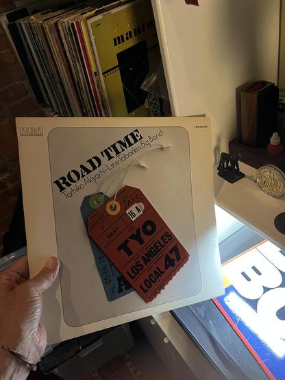 This photo shows [me] holding a vintage album titled "ROAD TIME" by Toshiko Akiyoshi-Lew Tabackin Big Band, released on RCA (CPL2-2242 STEREO) as a 2-record set. The cream-colored album cover features a clever design with overlapping travel-themed luggage tags in different colors - including a red tag for "TYO LOS ANGELES LOCAL 47" and blue/green tags with flight numbers.

The setting shows a vinyl collection with records stored vertically in the background. A yellow album (possibly "Mañana" by…