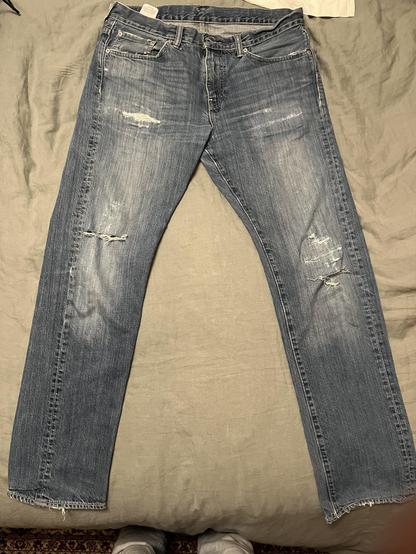 This photo shows a pair of well-worn, distressed blue jeans laid flat on a beige bedspread. The jeans are positioned with the front facing up, displaying classic denim features including:

A medium blue wash with significant fading and wear patterns

Multiple distressed areas and rips, particularly visible on both thighs and the right knee area

Natural whisker patterns and fading around the thighs and seat area

Traditional five-pocket styling with visible rivets

A button fly closure [I think…