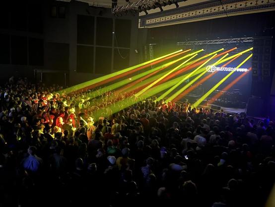 This photo captures an energetic live concert in a large indoor venue. The image shows a packed crowd of hundreds of attendees filling the floor space, with people standing densely together facing toward a stage area on the right side of the frame.

The lighting design is spectacular, featuring dramatic beams of colored light - primarily yellow, orange, and some red/pink tones - cutting diagonally across the venue from right to left. These sharp, parallel light beams create a dynamic visual eff…