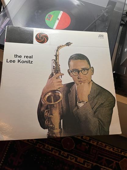 This photo shows a classic jazz album titled "the real Lee Konitz" on the Atlantic label (catalog number P-6234A). The album cover features a striking portrait-style photograph of saxophonist Lee Konitz wearing dark-framed glasses and a textured suit jacket. He's posed in a contemplative manner with his hand near his chin while holding his saxophone, which is prominently displayed in the frame.

The album is positioned in front of a turntable, with a vinyl record visible on the platter behind i…