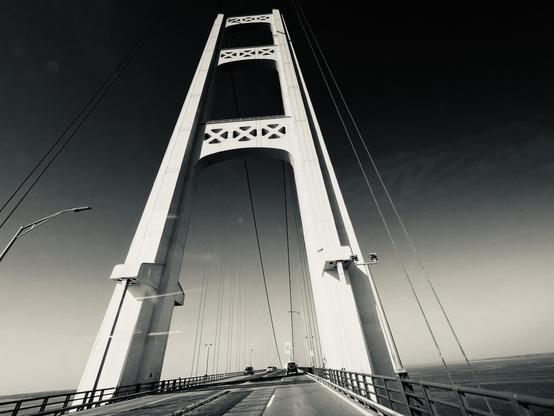 This black-and-white photo shows a striking view of a large suspension bridge, photographed from the roadway as a vehicle travels across it. The image prominently features one of the bridge’s tall towers, with its geometric cross-bracing details visible near the top. Thick suspension cables rise gracefully from both sides of the deck, converging at the tower and extending into the distance toward the next span.

The composition emphasizes the height and symmetry of the structure, with a dramati…