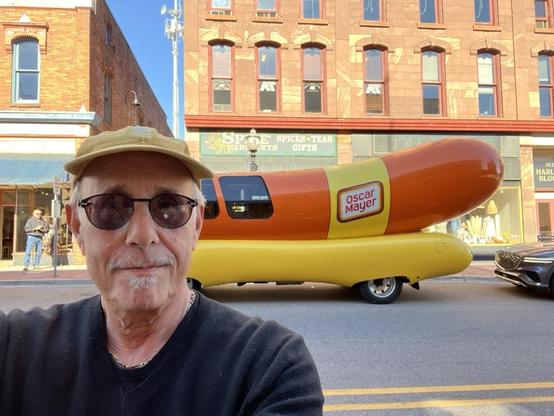 I am taking a selfie on a sunny street in Marquette, MI. Behind me is the iconic Oscar Mayer Wienermobile — a large, hot-dog-shaped vehicle painted in yellow and orange with the Oscar Mayer logo on the side. The scene is in a small downtown area with historic brick buildings housing shops; one storefront sign reads “Spices • Teas • Gifts.” I am wearing a tan cap, sunglasses, and a black sweater, and am smiling slightly at the camera.