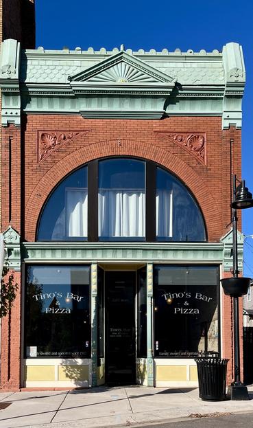 The photo shows the facade of a brick building that houses a business named Tino's Bar & Pizza. The building has a prominent arched window on the second floor and a green, decorative cornice at the top. The ground floor features two large windows displaying the business name and a central entrance. The text on the windows indicates that the business is "Family owned and operated since 1982". The image was taken on a clear, sunny day.