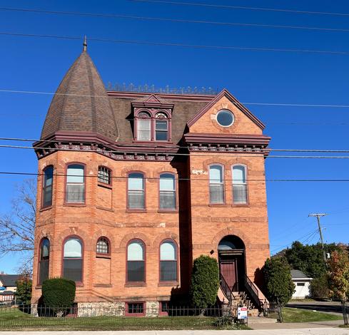 The photo shows a historic, three-story red brick building with a prominent turret, located at 101 S Pioneer Avenue in Negaunee, MI. The building features arched windows and a dark-colored roof. The property was originally built as a home and later converted to office space. 