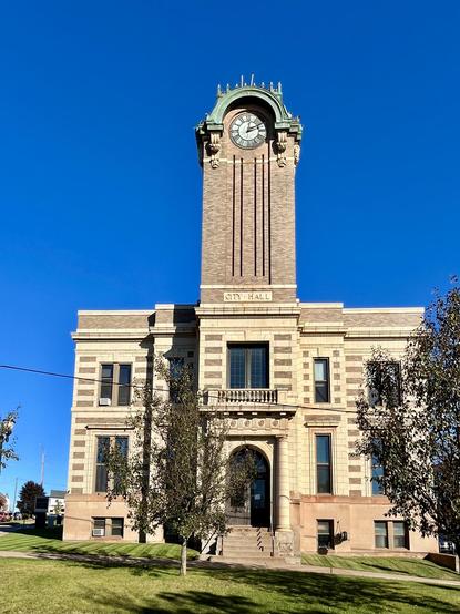 The photo shows the Negaunee City Hall, a historic two-story building in Negaunee, Michigan, with a prominent clock tower. 

Architecture: The building, erected in 1915, is constructed with alternating courses of white pressed brick and grayish-white limestone, featuring terra-cotta detailing. 

Clock Tower: A ninety-four-foot-high clock tower rises above the main entrance. 

Location: The building is located in Negaunee, Michigan. The words "CITY HALL" are visible above the main entrance. 