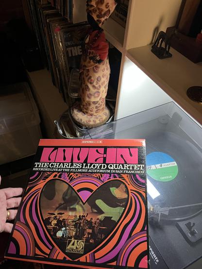 This photo captures a vinyl record collection scene with several interesting elements:

Main focus: A Charles Lloyd Quartet album - "Recorded Live at the Fillmore Auditorium in San Francisco" on Atlantic Records. The album cover features vibrant psychedelic artwork typical of the late 1960s, with swirling orange, pink, and purple patterns framing a photo of the band performing on stage.

Turntable setup: The record is being held over a modern turntable with a clear dust cover. The turntable sit…