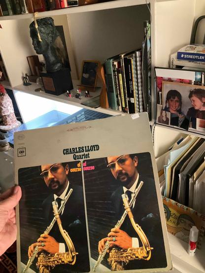 This photo reveals a Charles Lloyd album from an impressive collection [oh please], shown in a personal, lived-in creative space.

The Album: "Of Course, Of Course" by the Charles Lloyd Quartet - featuring a striking cover design with a split image showing Charles Lloyd in formal attire (white shirt, bow tie, and dark suit) holding his flute. The dual image creates a mirrored effect, with colorful "of course" text in orange and pink between the two portraits. Lloyd wears sunglasses and is photo…
