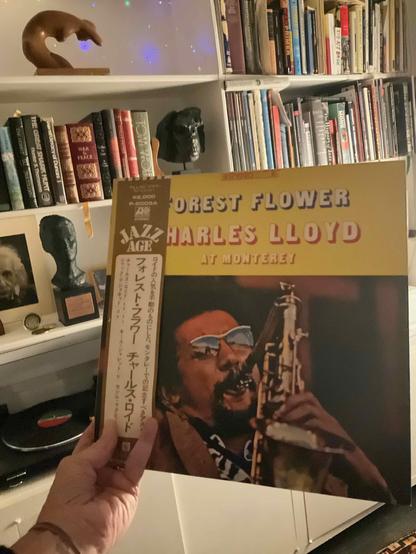 This photo showcases a rare Japanese pressing of a Charles Lloyd album, displayed in a record collector's library.

The Album: "Forest Flower" by Charles Lloyd - specifically a Japanese pressing on the Atlantic Jazz Age label. The cover features a golden-yellow background with typography displaying "FOREST FLOWER" and "CHARLES LLOYD" in red and cream lettering, with "AT MONTEREY" below. The main image shows Charles Lloyd wearing sunglasses and a cap, captured mid-performance playing his saxopho…
