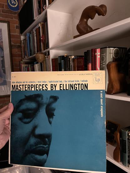 This photo showcases a classic Duke Ellington album from a home library.

The Album: "Masterpieces by Ellington" on Columbia Records - a stunning vintage LP with one of the most iconic album covers in jazz history. The design features a dramatic close-up portrait of Duke Ellington rendered in deep teal/turquoise tones against a cream-colored upper section. The bold black typography of "MASTERPIECES BY ELLINGTON" dominates the top, with track listings including "Mood Indigo," "Sophisticated Lady…