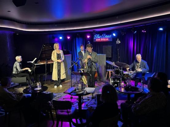 This photo captures a live jazz performance at the Blue Llama Jazz Club in Ann Arbor last night. The intimate venue features the Ellen Rowe Quartet ft. Mike Sakash and Sunny Wilkinson performing on stage, bathed in atmospheric purple and blue lighting that creates a classic jazz club ambiance.

The ensemble consists of five musicians: pianist Ellen Rowe at a grand piano on the left, vocalist Sunny Wilkinson in an elegant gold/yellow dress with black accents standing center-left, Rob Bickley on …