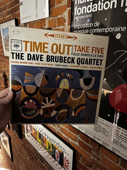 My hand is holding up a classic vinyl record album: "Time Out" by The Dave Brubeck Quartet, Columbia Records 1959 stereo release (catalog number CS 8192), one of the most celebrated jazz albums of all time and one of the first jazz albums to sell over a million copies. It features the classic lineup of the quartet: Dave Brubeck on piano, Paul Desmond on alto saxophone, Joe Morello on drums, and Eugene Wright on bass. The track listing visible includes "Strange Meadow Lark," "Three to Get Ready,…