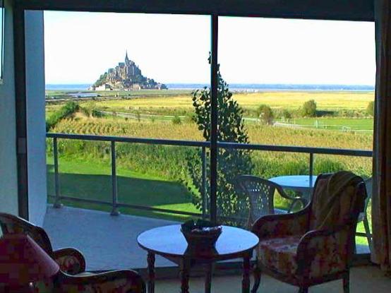 This photograph captures a stunning view from inside a hotel room looking out through large glass doors onto a balcony or terrace. The view showcases Mont-Saint-Michel, the famous medieval abbey and fortress situated on a tidal island in Normandy, France.

In the foreground, there are two floral-patterned upholstered chairs flanking a small round dark table, creating a cozy seating area. The room opens onto a balcony with glass railings and what appears to be outdoor furniture including a blue …