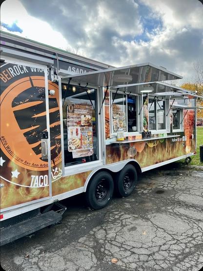 This photo shows a vibrant food truck with eye-catching branding. The truck features a distinctive design with flame graphics wrapping around the exterior, giving it an energetic, fiery aesthetic. The prominent circular logo displays what appears to be tacos and includes text mentioning “ASADA” and location information for Ann Arbor.

The truck has a modern design with large serving windows that fold open, allowing customers to order from outside. A menu board is visible on the side, and there’…