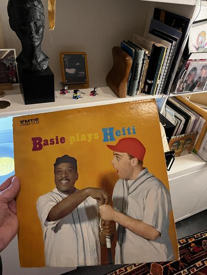 I am holding up a vinyl record album with a bright yellow/orange cover. The album features Count Basie and Neal Hefti dressed in vintage baseball uniforms - one wearing a dark cap and the other wearing a red cap. The text on the cover reads "Basie plays Hefti" (with "EMUS" visible at the top, part of a reissue label).

The photo is taken in a home workspace. In the background, there's a white shelving unit containing various items including:

• A black sculptural bust on the left
• A small fram…