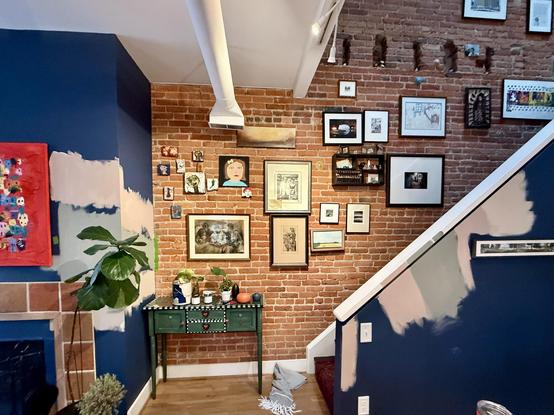 This photograph shows a stylish, eclectic interior space with high ceilings and exposed brick walls. The room features a dramatic gallery wall covering most of the brick surface, filled with an assortment of framed artwork, photographs, and decorative pieces in various sizes and styles.

The architectural details include white exposed beams on the ceiling and a white support beam. A staircase with white railings is visible on the right side of the image, ascending at an angle.

The walls are pa…