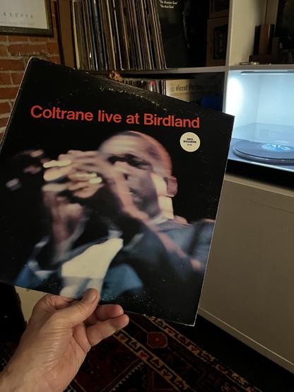 I am holding up a vinyl record album titled "Coltrane live at Birdland" in red text against a black background. The cover features a dynamic, slightly blurred performance photograph of John Coltrane playing saxophone, captured in motion with his hands visible on the instrument. He is wearing a dark suit. 

The photo is taken in a home music collection space, with extensive vinyl record storage visible in the background - tall shelves packed with LP spines can be seen. There's also exposed brick…