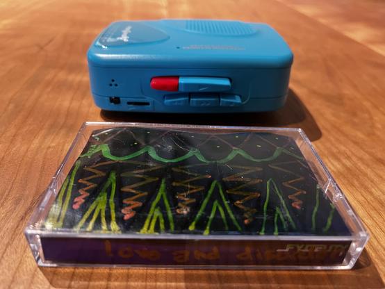 This photograph shows a portable cassette player positioned on a table, with a cassette tape displayed in front of it.

The cassette player has a distinctive teal color and features a prominent red power/play button on its front panel, along with playback control buttons. There's also a small speaker grille visible on the top of the device, and various ports and controls on the front face.

In front of the player sits a cassette tape in a clear plastic case. The tape features striking artwork w…