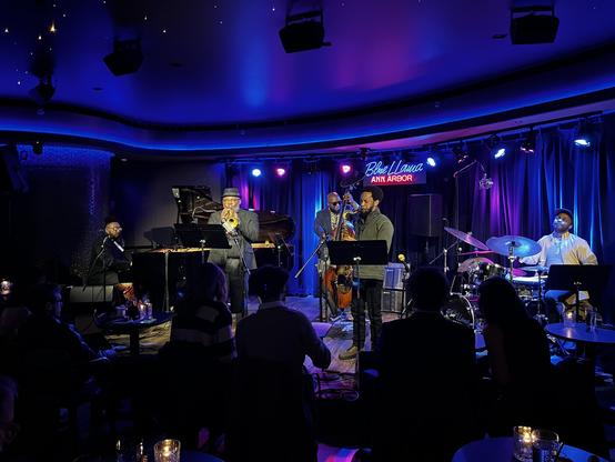 This is a captivating photograph of a live jazz performance at the Blue LLama Ann Arbor venue. The intimate club setting features a jazz quintet performing on stage, with moody blue and purple stage lighting creating a classic jazz club atmosphere.

The ensemble includes a pianist at a grand piano on the left, Dwight Adams on trumpet in the center-left wearing a fedora and suit, an upright bassist in the center-back, marcus Elliot on sax in the center reading from sheet music, and a drummer on …
