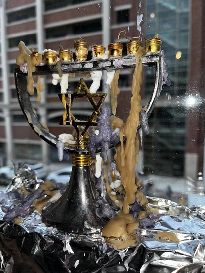This photo shows a menorah (a nine-branched Jewish candelabrum) that has been used for Hanukkah celebrations. The menorah is made of brass with gold-colored cups for holding candles. 

What’s particularly striking about this image is the dramatic aftermath of use - the menorah is covered in extensive wax drippings that have accumulated and hardened in flowing, organic shapes. The wax appears in shades of cream, tan, and purple-gray, creating almost sculptural formations that cascade down the br…