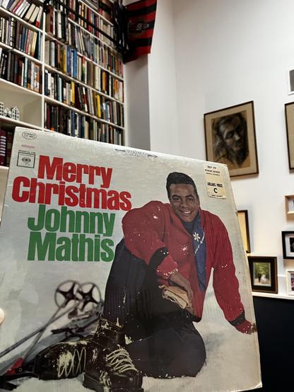This photo shows a vintage "Merry Christmas" album by Johnny Mathis being held up in a cozy home library. The album cover features the legendary singer in a festive red cardigan sweater, sitting in a snowy setting with a warm, cheerful smile. The title is displayed in bold red and green lettering, perfectly capturing the holiday spirit.

The background reveals an floor-to-ceiling bookshelf packed with books. White walls display several framed artworks, including a portrait and other pieces in v…