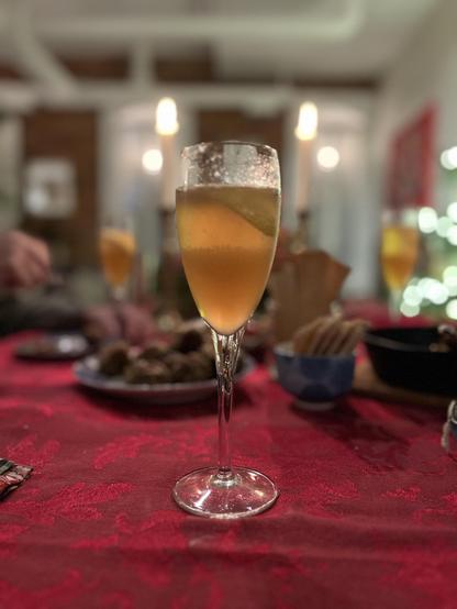 This photo captures an elegant champagne flute positioned on a festive dining table covered with a rich red tablecloth featuring a decorative pattern. The glass contains a golden-hued bubbly beverage.

The setting suggests a holiday gathering. In the soft-focused background, you can see warm, glowing candles creating ambient lighting, plates of food, and other dining elements that contribute to an intimate, festive atmosphere. The composition centers the champagne flute in sharp focus while the…