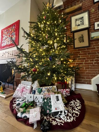 This photo shows a beautifully decorated Christmas tree in a modern home with exposed brick walls and hardwood floors. The tree is a full, natural-looking evergreen adorned with warm white string lights that create a soft, inviting glow throughout its branches.

The tree features a mix of ornaments including a gold star topper, colorful glass baubles (red, blue, and other colors), and elegant decorative pieces like white origami-style ornaments and angel figures. The decorating style is eclecti…