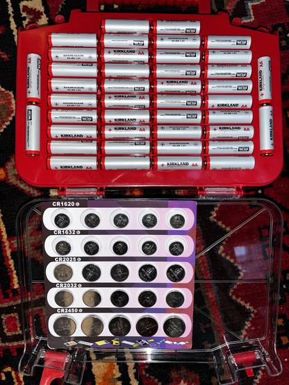 This photo shows an even more detailed view of a well-organized battery storage system, photographed from above on the same beautiful Persian rug.

Top Section - Red Battery Organizer:

This contains multiple rows of Kirkland brand AA batteries (alkaline, 1.5V) neatly arranged in a red plastic storage case. The white cylindrical batteries are systematically placed in horizontal rows with their positive and negative terminals clearly visible and marked. There appear to be approximately 40+ AA ba…
