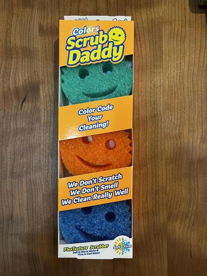 This photo shows a package of "Colors Scrub Daddy" cleaning sponges displayed on a wooden surface. The bright, eye-catching packaging features the cheerful Scrub Daddy brand design with its signature smiley face logo.

Package Contents:

The clear window display shows three colorful sponges stacked vertically:

Teal/turquoise sponge (top) with a smiling face
Orange sponge (middle) with a smiling face
Blue sponge (bottom) with a smiling face

Each sponge has the characteristic Scrub Daddy design…