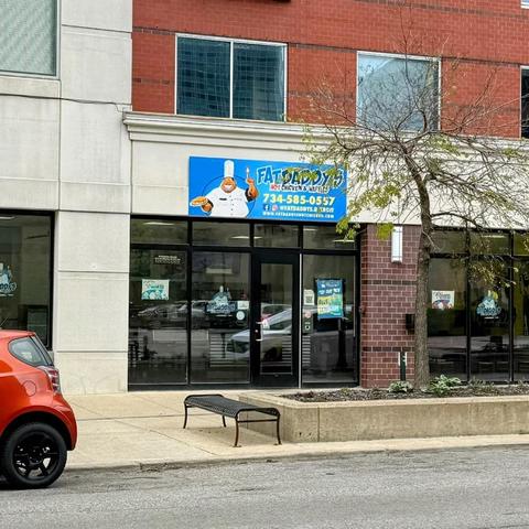 This photo shows the storefront of "Fat Daddy's Hot Chicken & Waffles," a restaurant located in Ann Arbor.

Building and Location:
The restaurant occupies a ground-floor retail space with contemporary architecture featuring a cream/beige upper facade and brown brick accent column. Large floor-to-ceiling black-framed glass windows and doors provide visibility into the interior. The building has additional floors above with red brick exterior and blue-tinted windows.

Signage and Branding:
A prom…