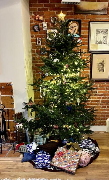 This is a cozy Christmas scene featuring a decorated evergreen tree in a home with exposed brick walls. The tree is adorned with white lights and various ornaments including eclectic handmade decorations. A gold star tops the tree.

The setting has a warm, artistic feel with the rustic red brick wall as a backdrop. Several framed artworks and photographs are hung on the brick, including be vintage photographs and portrait sketches. There are also some smaller photos or artwork pieces attached d…