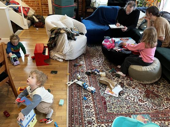 This photo captures a lively family moment in a cozy loft-style living room with exposed brick walls and hardwood floors.

The Space:
A warm living area featuring a large ornate Persian rug in burgundy, cream, and navy patterns. The room has an eclectic, bohemian feel with mixed furniture and exposed brick.

The People:

Left: A child in a colorful striped shirt sits with a toy truck
Foreground: A toddler in a white vest and gray shirt holds a toy, looking upward
Right side: An adult in black s…