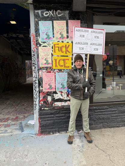 I'm standing by Trapper's Alley next to a wall covered in small posters. I'm dressed in cold-weather clothing including a dark jacket, beige pants, hiking boots, and a knit cap, and am holding a protest sign.

The sign reads "ABOLISH ICE" repeated four times in a grid pattern, with illustrations of melting ice cubes. There's also a bright yellow sign attached to the wall behind me that says "FUCK ICE" in bold red letters.

The wall itself is heavily postered with multiple flyers advertising wha…
