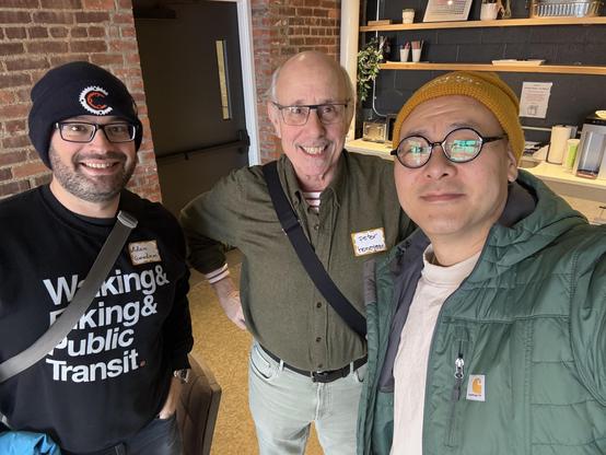 This photo shows Adam Goodman, me, and Dug Song posing together for a selfie in a casual office setting with exposed brick walls and wooden shelving in the background.

On the left, Adam is wearing a black beanie with a logo, glasses, and a black t-shirt that reads "Walking & Biking & Public Transit." He has a beard and is wearing a messenger bag strap across his chest. His name tag reads "Adam Goodman."

In the center is an older gentleman with glasses and a warm smile, wearing an olive green …