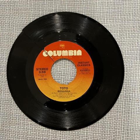 [describe this photo]

This photo shows a vintage 7-inch vinyl record (a 45 RPM single) resting on a textured burlap or linen fabric background. The record is on the Columbia Records label, identifiable by its distinctive orange and yellow gradient label design with the Columbia logo at the top.

The label indicates this is "Rosanna" by Toto, released in March 1982 as part of Columbia's "Instant Classics" series. The record shows it's a stereo recording with a duration of 3:59. You can see the …