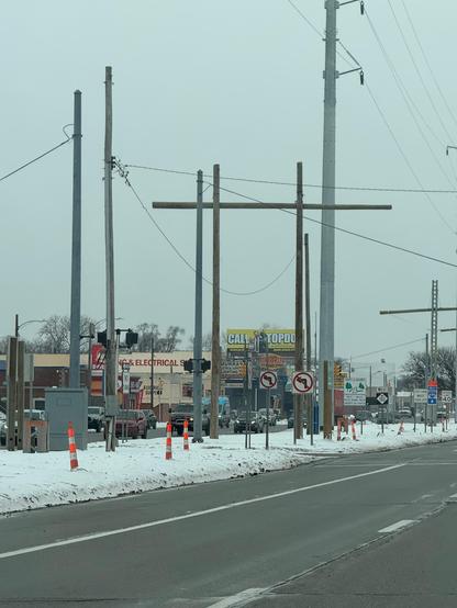 This photo shows a winter street scene with snow on the ground. The main focus is on utility infrastructure - there are several tall utility poles with power lines running between them creating a web of cables across the cloudy gray sky. In the center, there's a distinctive H-frame utility structure made of tall wooden or concrete poles with a horizontal crossbeam.

The road appears to be clear and wet, with white lane markings visible. Orange and white construction cones line the side of the r…