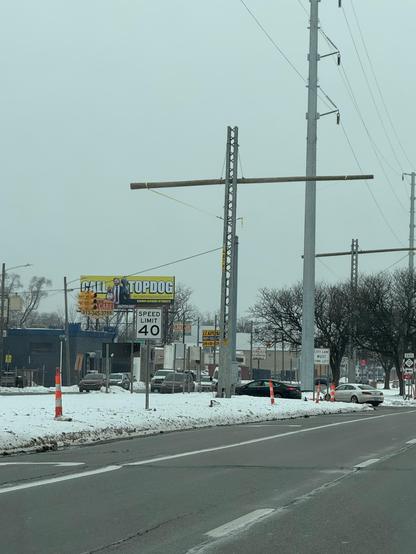 This photo shows another view of a similar winter street scene. The image features tall utility poles with H-frame crossbeams supporting power lines against an overcast gray sky. The poles appear to be metal lattice or truss-style construction with horizontal crossbeams.

In the foreground, there's a clear road with white lane markings and a speed limit sign showing 40 mph. Snow covers the ground on both sides of the road, and orange construction cones are visible along the roadside. Several ve…