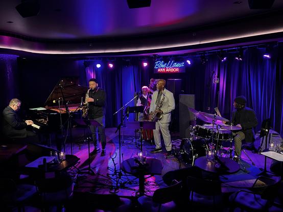 This is a beautiful shot of a jazz performance at the Blue LLama in Ann Arbor! The venue has fantastic ambiance with its intimate stage, purple and blue mood lighting, and draped curtains.

The quintet is mid-performance with:
	∙	A pianist on the left
	∙	Two saxophonists (one playing a tenor sax in darker clothing, another in a light-colored suit)
	∙	A bass player in the back
	∙	A drummer on the right

The stage lighting creates a wonderful atmosphere, and you can see music stands suggesting th…