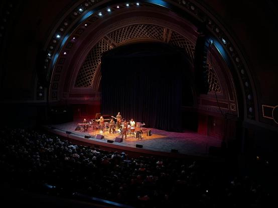 This photo captures jazz legends Terence Blanchard and Ravi Coltrane performing with The E-Collective at the iconic Hill Auditorium in Ann Arbor, Michigan.

The Performance:
Terence Blanchard (renowned trumpeter and composer) and Ravi Coltrane (acclaimed saxophonist and son of John Coltrane) are performing with The E-Collective, Blanchard's electric fusion ensemble

The intimate stage setup features the musicians arranged with their instruments under focused stage lighting

The warm glow create…