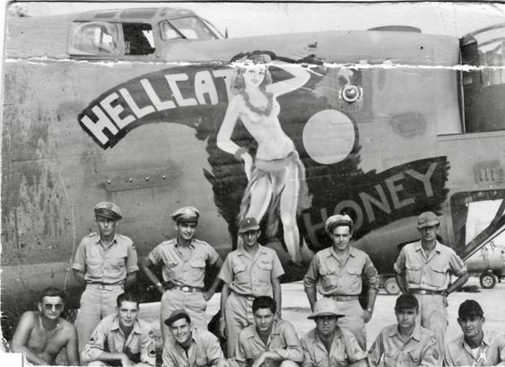 This is a black and white photograph from World War II showing an aircrew posing in front of their B-24 Liberator heavy bomber.

The Aircraft:
A B-24 Liberator bomber with distinctive nose art featuring a pin-up girl and the name "HELLCAT HONEY" prominently displayed

The aircraft shows visible battle damage and wear, indicating it has seen combat missions

The nose art depicts a classic 1940s-style pin-up girl in a patriotic pose wearing a lei and a grass skirt, typical of WWII bomber decorati…