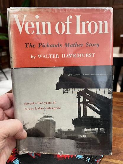 This photo shows a vintage hardcover book with its dust jacket titled "Vein of Iron" with the subtitle "The Pickands Mather Story" by Walter Havighurst.

Book Details:
The dust jacket features a coral/salmon pink upper portion with white lettering

Below is a black and white photograph showing an industrial Great Lakes scene

The photograph depicts a cargo ship (ore carrier) passing under a large industrial loading dock or bridge structure

The subtitle reads "Seventy-five years of Great Lakes …