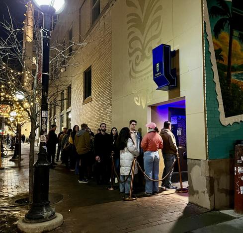 This nighttime photo captures a line of people waiting outside Huna, a trendy establishment in downtown Ann Arbor.

The Scene:
A queue of approximately 15-20 young adults dressed casually for an evening out waiting along a brick sidewalk

A velvet rope barrier controls the line near the entrance

The Venue:
The building features a distinctive cream-colored facade with subtle tropical-inspired relief designs

A prominent blue illuminated sign reading "HUNA" with a pineapple logo

Purple/blue atm…